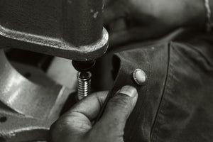 A close-up black and white photo of a hand pressing a metal snap button onto a stac jacket at the manufacturer in the UK. 