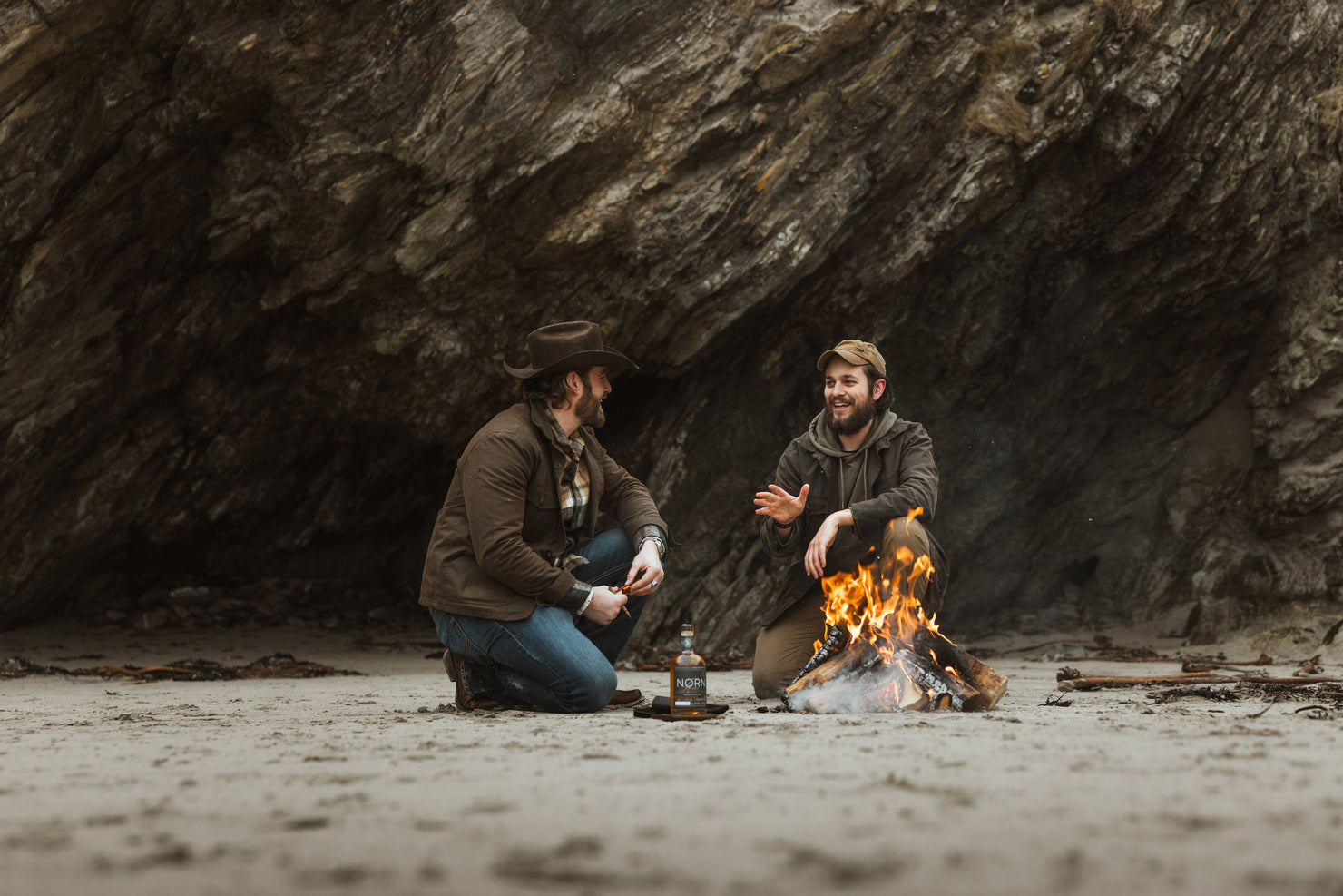 Bear&Wolf founders Jake and Bill in conversation around a beach campfire in Shetland, both wearing Cairn Overshirts in Raven with Bill in a Wax Cap.