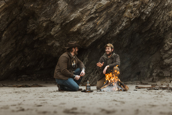 Bear&Wolf founders Jake and Bill in conversation around a beach campfire in Shetland, both wearing Cairn Overshirts in Raven with Bill in a Wax Cap.
