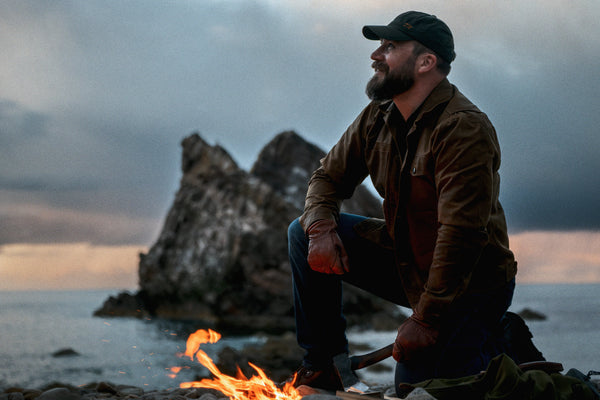 Man wearing the Stac in Saddle Brown and a Wax Cap, kneeling by a campfire below a sea stack at dusk.