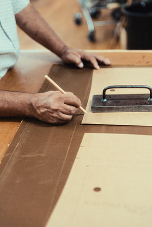 A close-up of a person's hands marking Halley Stevensons waxed cotton with a pencil, using a card template on a workbench to make a saddle brown Stac jacket.