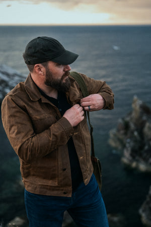 A man wearing the Stac in Saddle Brown and Wax cap walking along the Scottish coastline at dusk. 
