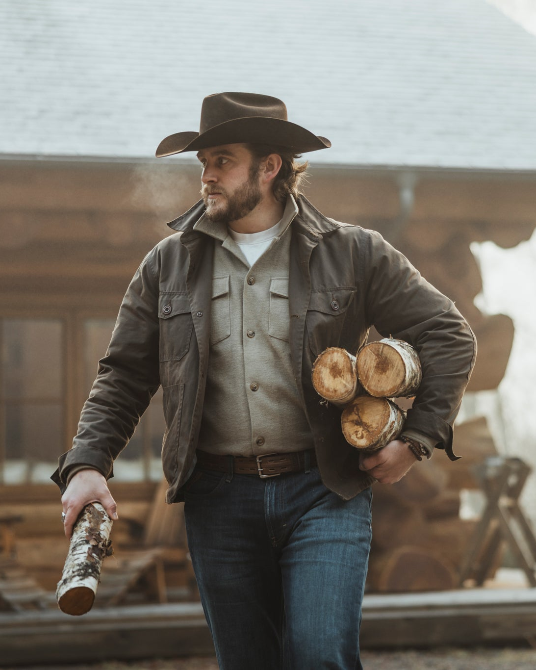 Bear&Wolf founder Jake wearing the Cairn Overshirt in Raven carrying chopped wood at a Scottish log cabin.