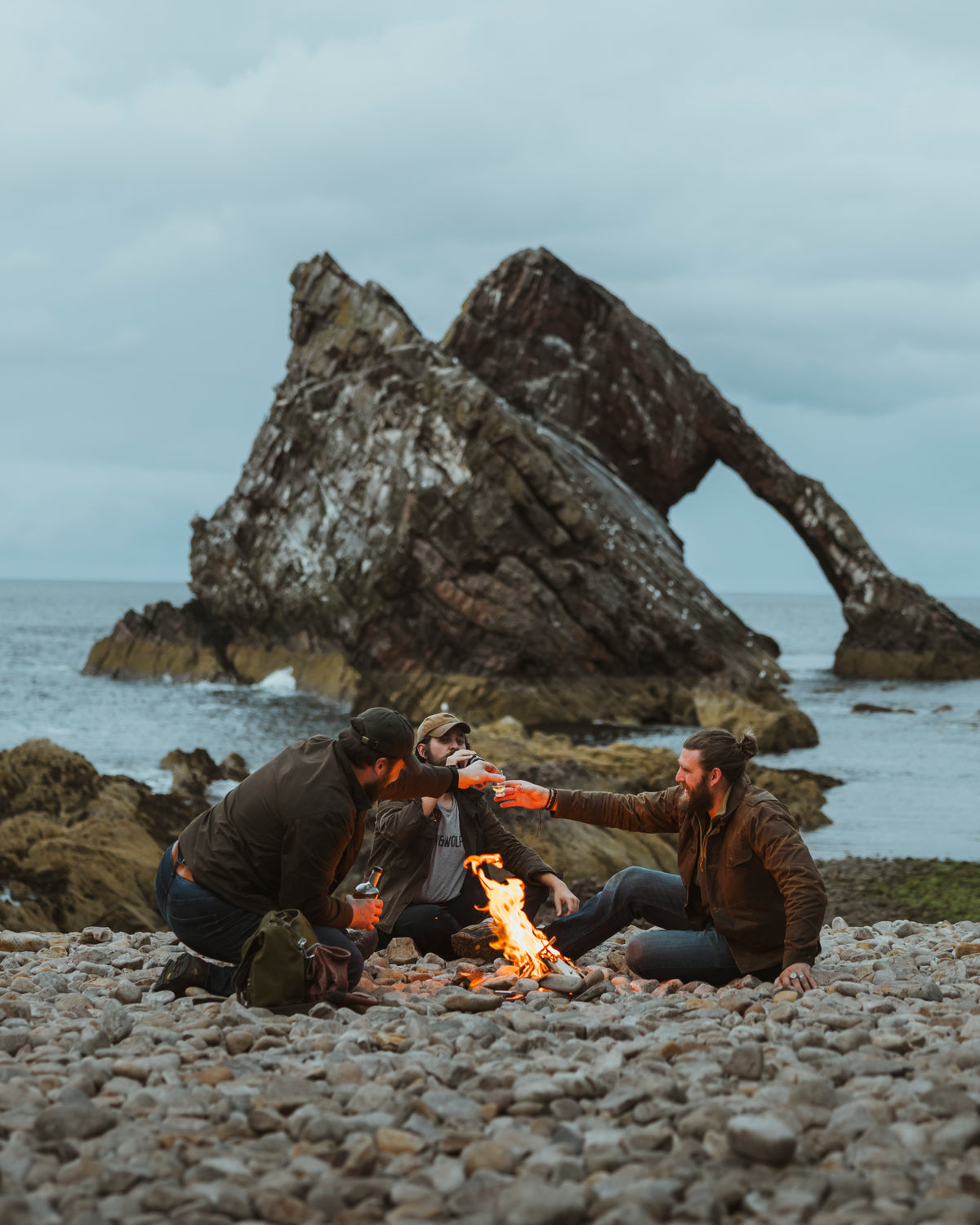 The founders of Bear&Wolf with a friend of the brand drinking whisky around a fire on the beach with Bow Fiddle rock behind them at sunset.