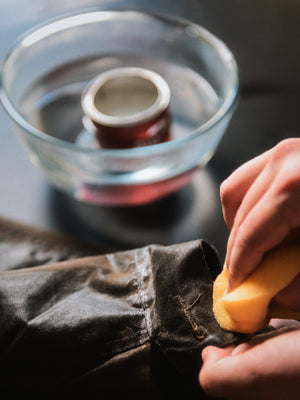 Applying Old Faithful No.01 wax treatment to the sleeve of a Cairn Overshirt using a sponge, with the open wax tin sitting in a bowl of hot water in the background.